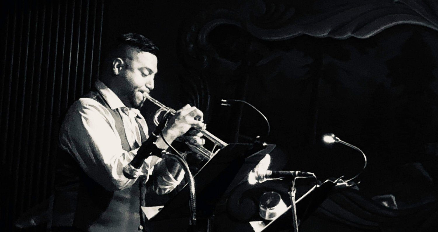 Black and white photo of Victor Garcia performing live on stage, playing trumpet under a spotlight. He wears a collared shirt and vest, standing in front of a music stand with mounted lights. A decorative mural is dimly visible in the background.