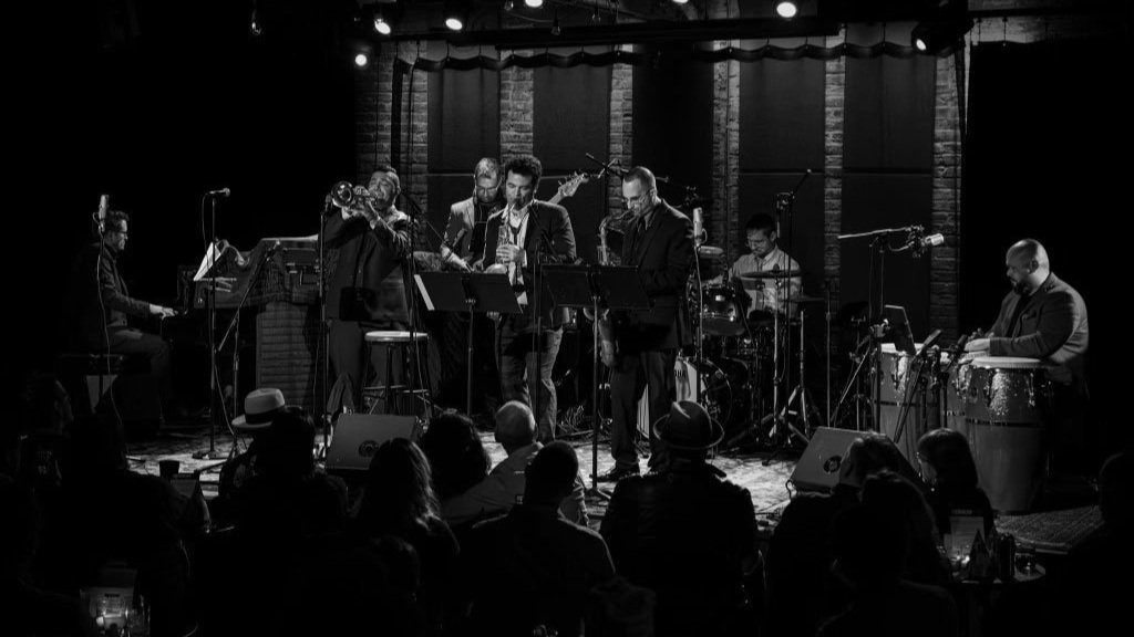 Black and white photo of a live jazz performance featuring an ensemble of eight musicians on stage. Victor Garcia plays trumpet near the center of the group. Other band members include a pianist, saxophonists, a drummer, a bassist, and a percussionist on congas. The audience is visible in silhouette in the foreground, watching the performance in a dimly lit venue.
