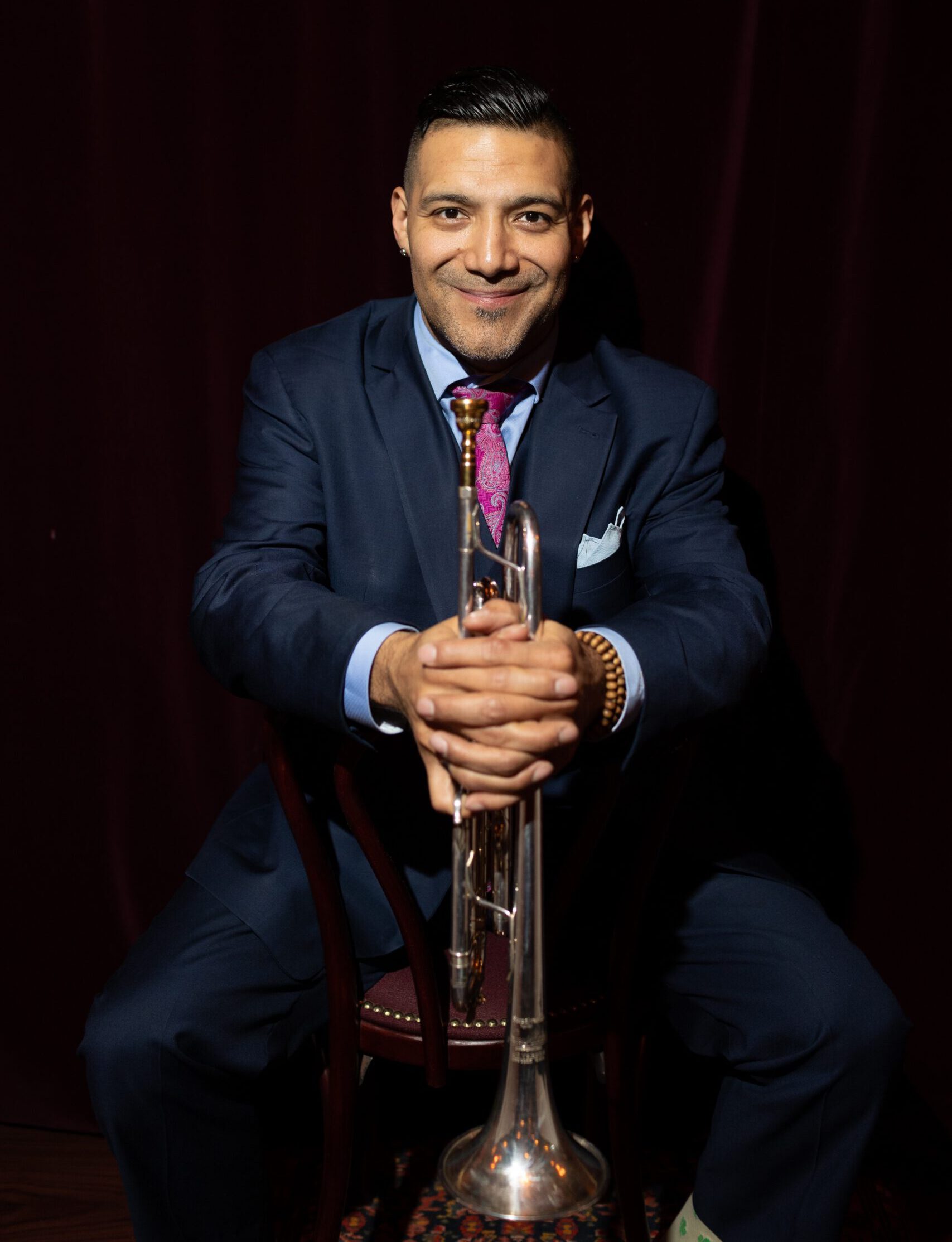 Jazz trumpeter Victor Garcia seated on a chair in a dark room, wearing a navy blue suit, a purple paisley tie, and a beaded bracelet. He is smiling at the camera while holding his trumpet vertically with both hands resting on the bell.