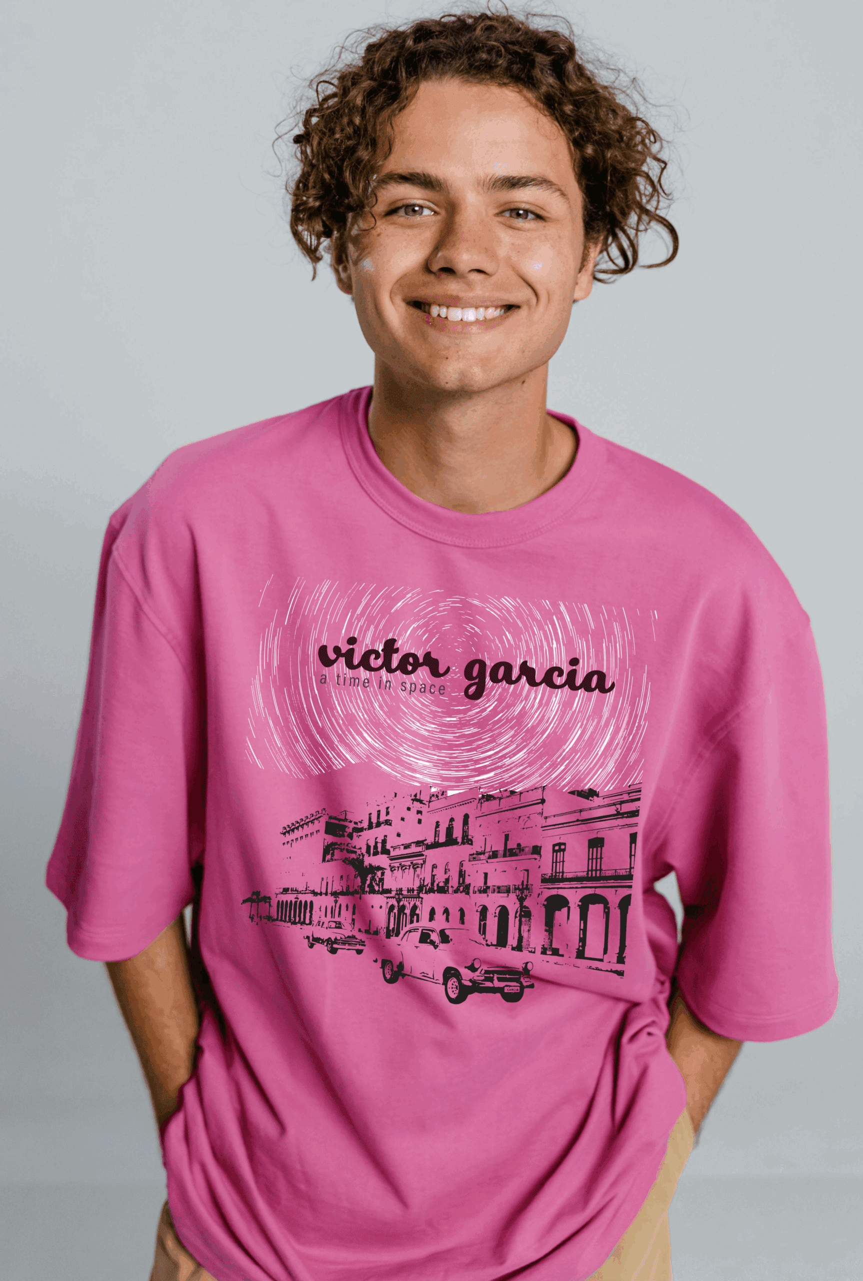 Smiling young man wearing a pink graphic t-shirt featuring the "Victor Garcia: A Time in Space" album design. The artwork includes swirling star trails above a cityscape of colorful colonial-style buildings and vintage cars, printed in black and white across the chest.